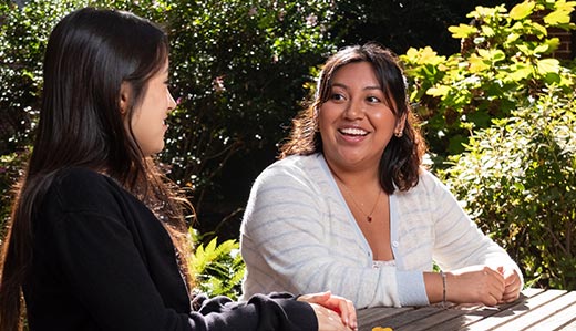 Two students sit at a wooden picnic table outside on Georgia Tech’s campus. The two students are having a conversation and smiling.